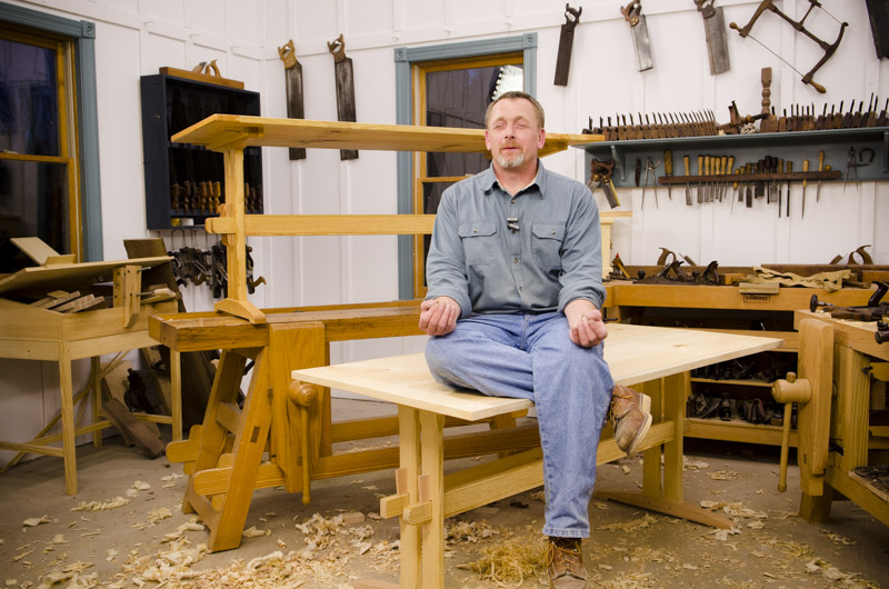 Will Myers sitting on the finished trestle table in a funny zen position next to woodworking workbenches in Joshua Farnsworth's woodworking workshop