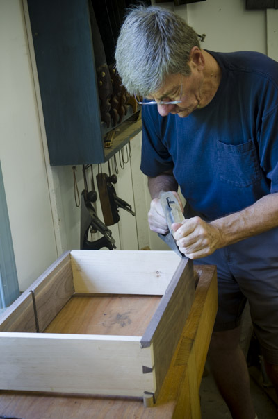 An older male student handplaning a walnut cupboard case with hand planes in the background