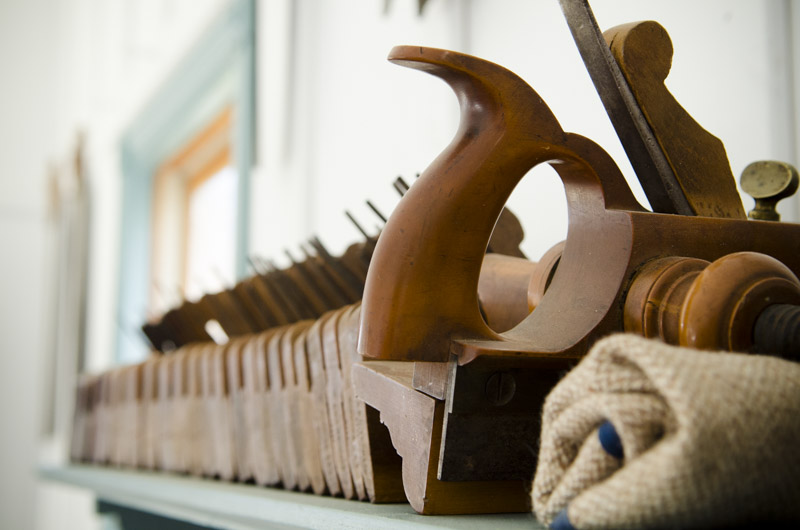 Antique hand planes and plow plane and moulding planes lined up on a blue hand tool shelf in Joshua Farnsworth's Wood And Shop Traditional Woodworking School